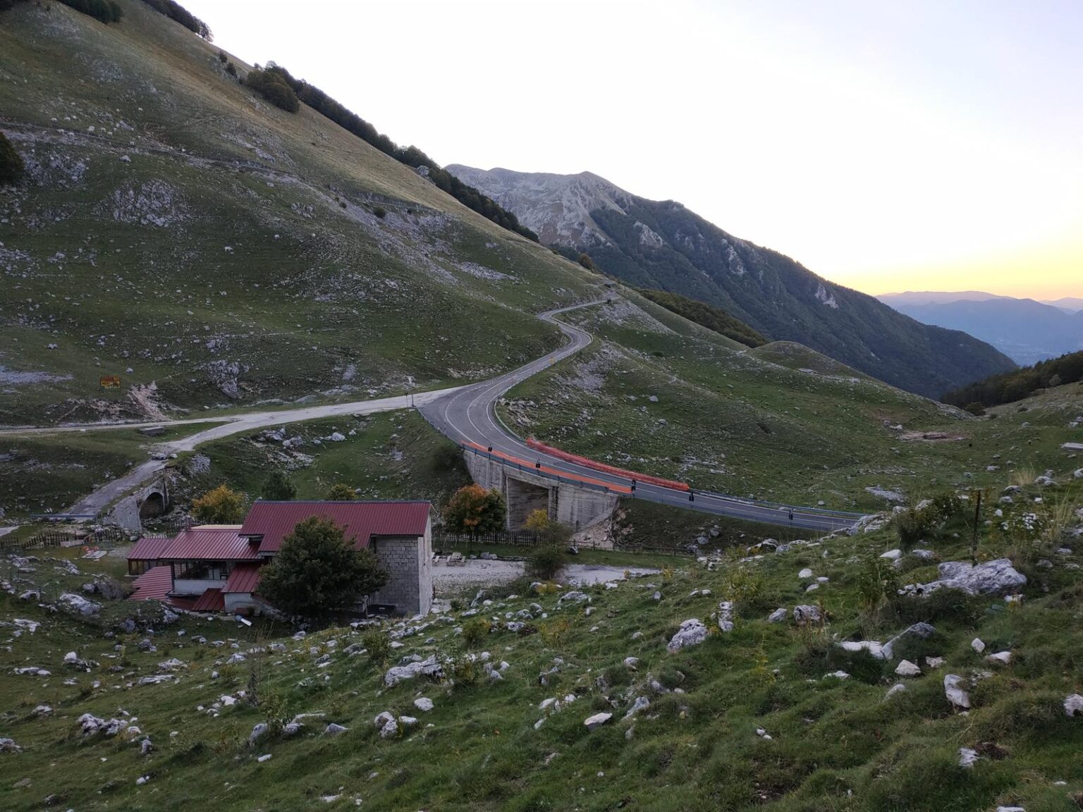 Valle con la strada del valico, un rifugio rosso e prati verdi al tramonto