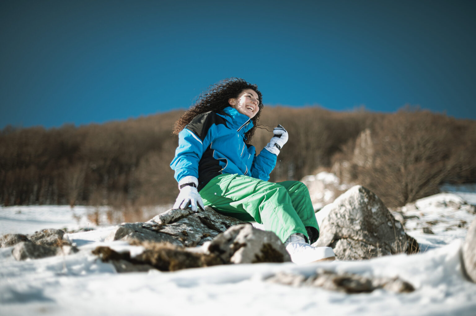 Escursionista seduta su una roccia nella neve, con giacca blu e pantaloni verdi, bosco spoglio sullo sfondo